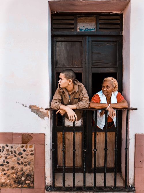 Free An adult and senior peering out from a rustic doorway with black railings. Urban setting, warm tones. Stock Photo