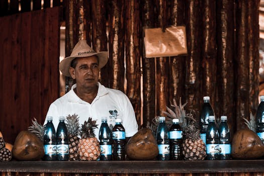 Man in a white shirt and hat selling bottled coconut and pineapple drinks at an indoor market.