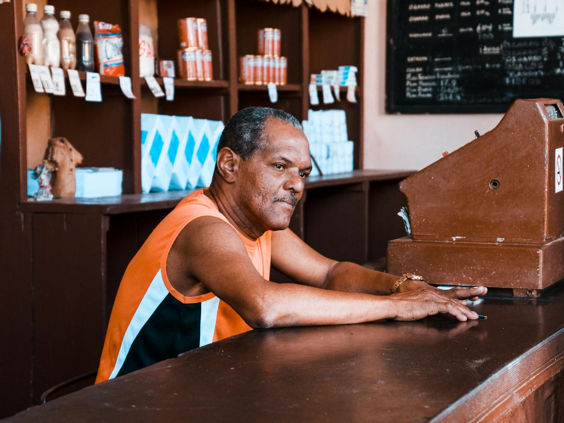 A man in a vintage setting, leaning on a wooden counter beside a cash register in a store.