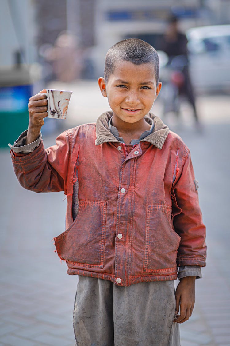 A Boy Wearing Torn Red Jacket Holding A Ceramic Mug