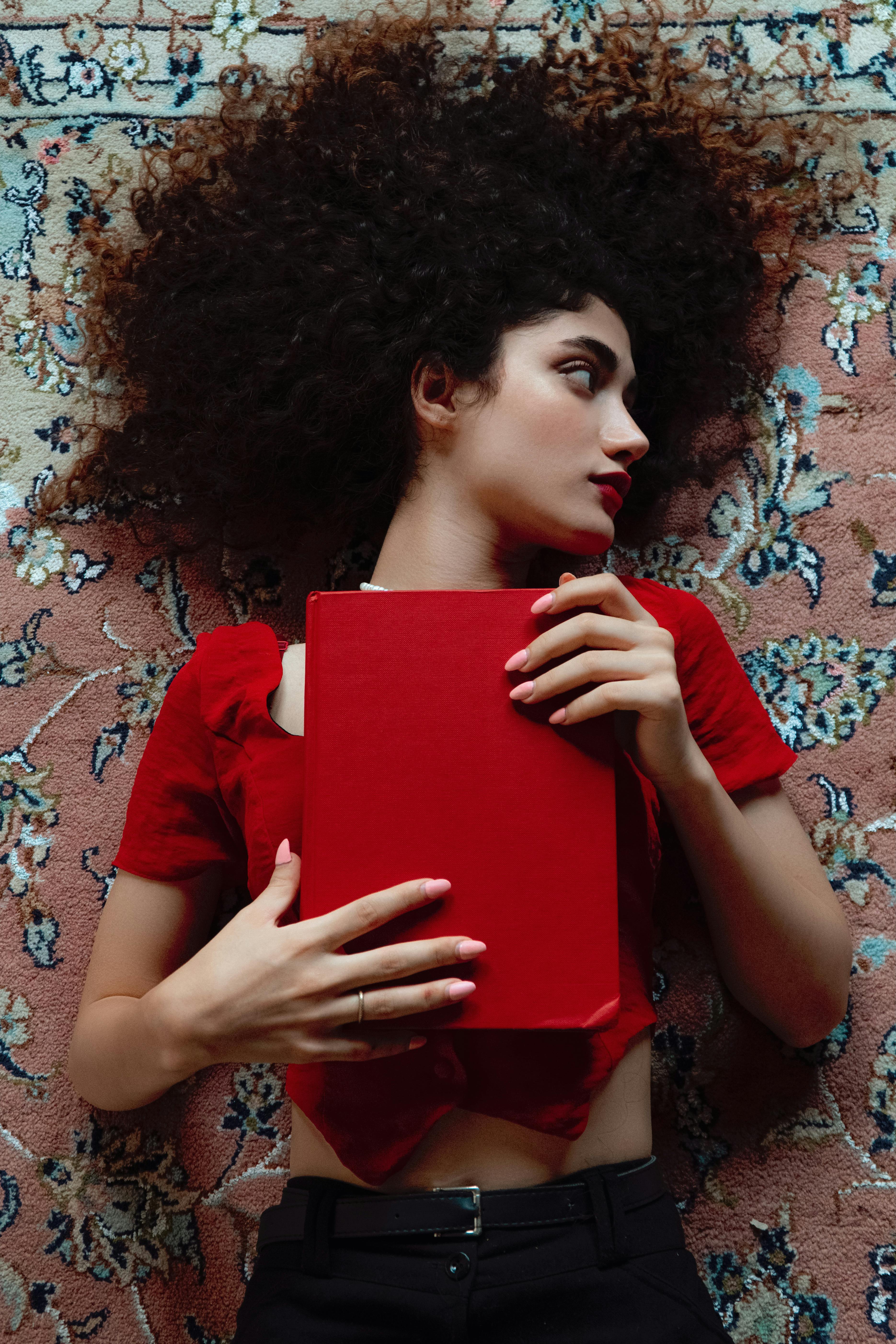 Woman with curly hair lying on carpet holding a red book. Indoor setting, artistic composition.