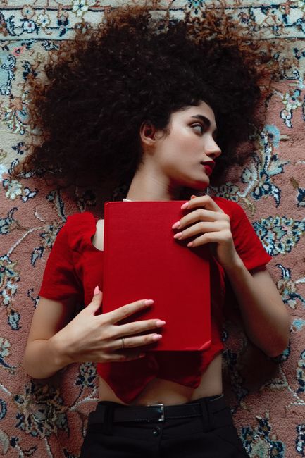 Woman with curly hair lying on carpet holding a red book. Indoor setting, artistic composition.