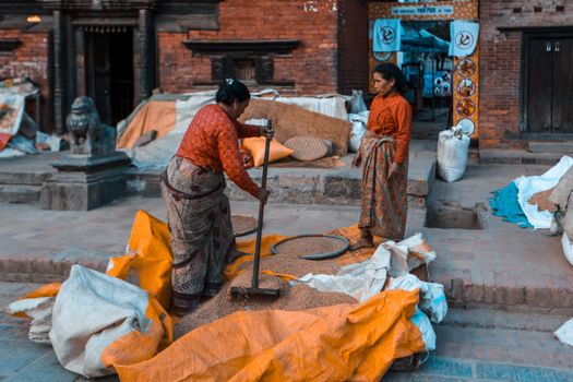Two women in traditional attire working on grains in an urban street scene.