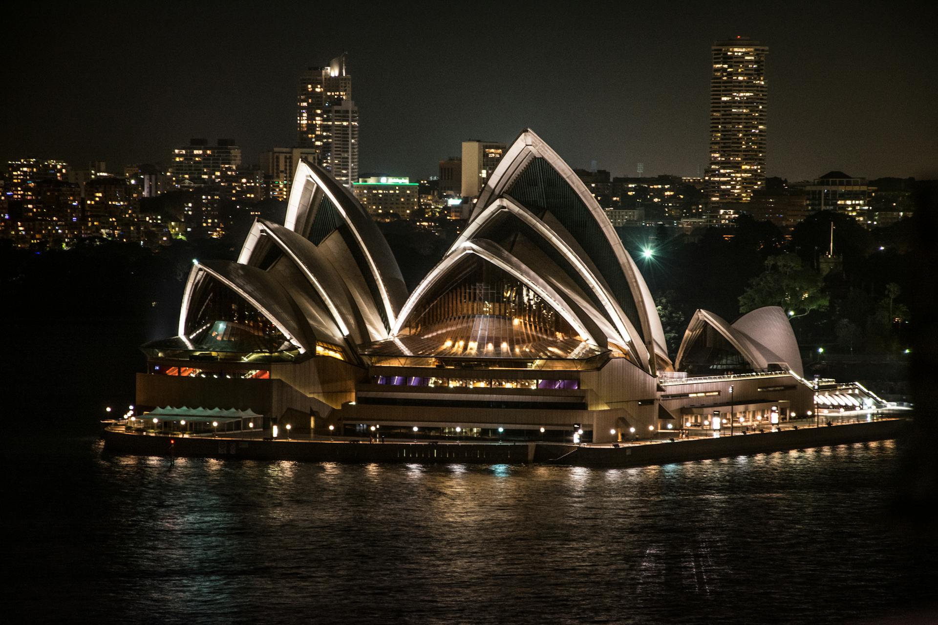 Illuminated Sydney Opera House on waterfront during nighttime, reflecting cityscape lights.
