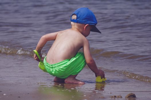 A young boy in bright shorts and cap playing on a sunny beach by the ocean waves.