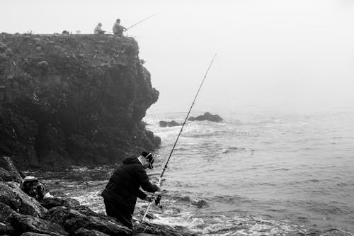 Free Grayscale Photo of a Man Fishing on a Rocky Shore Stock Photo
