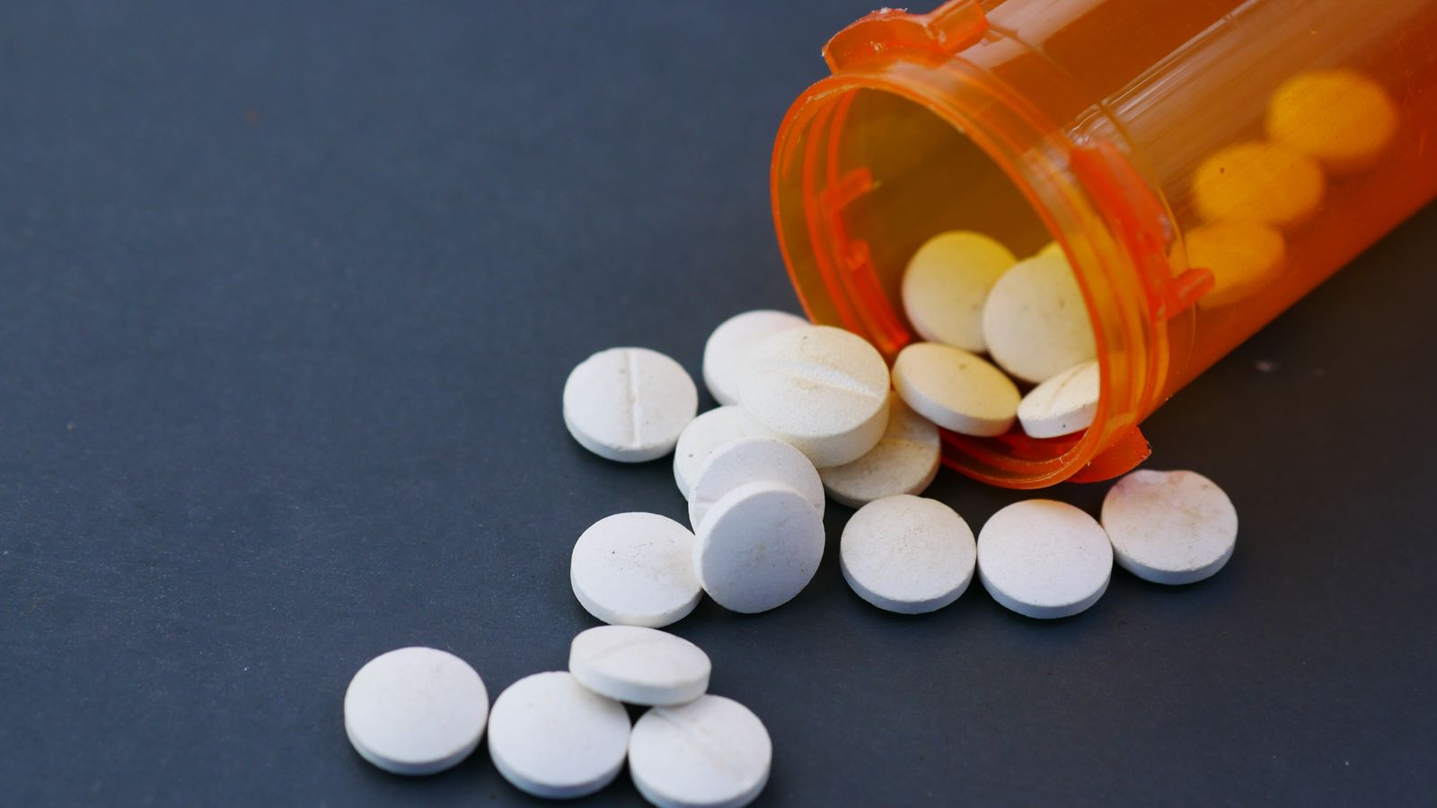 Close-up of white pills scattered from an orange prescription bottle on a gray surface.