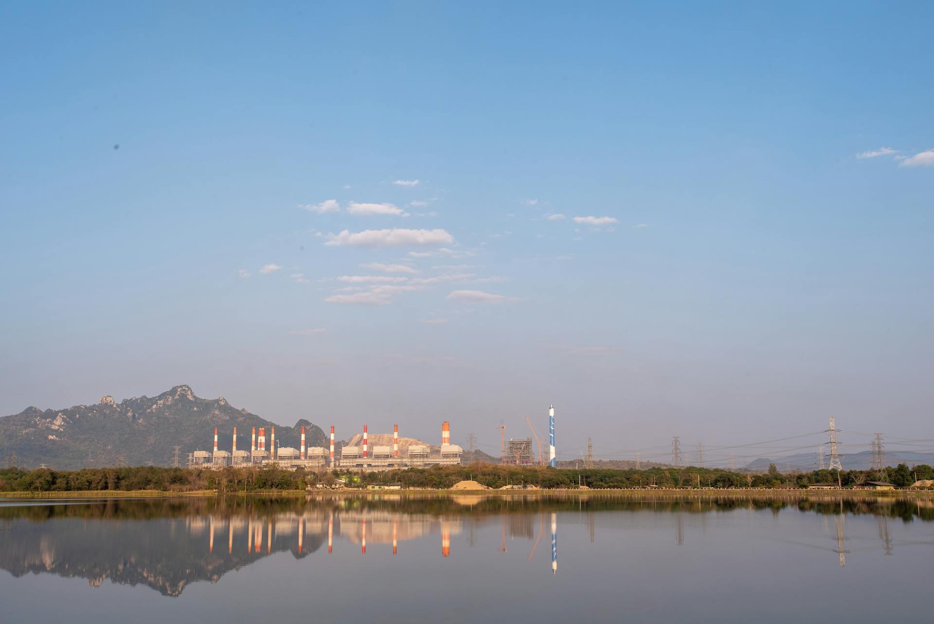 Scenic view of a power plant by a lake, reflecting mountains and a sky with scattered clouds.