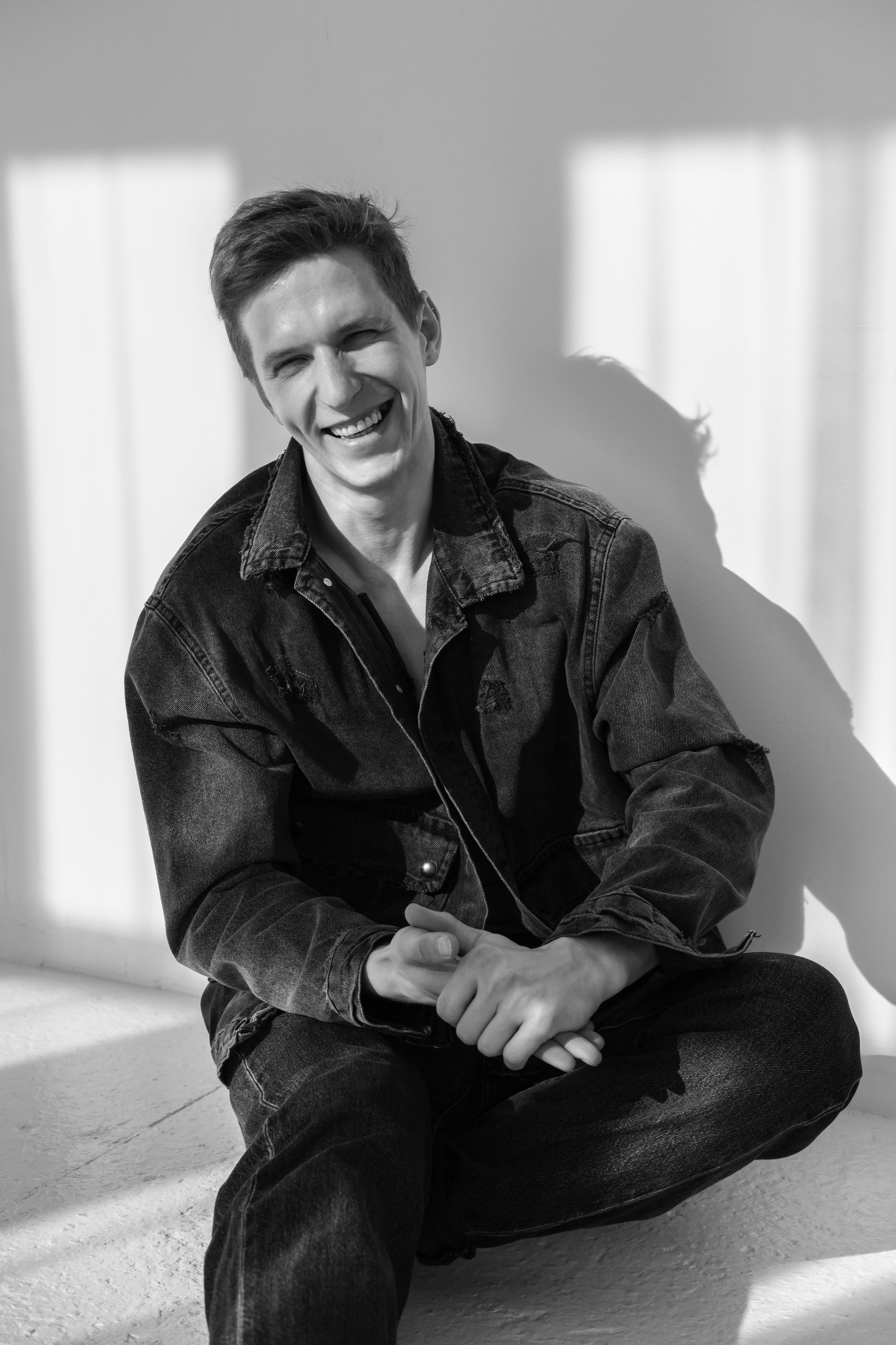 A cheerful man sits casually in a denim jacket, captured in grayscale.