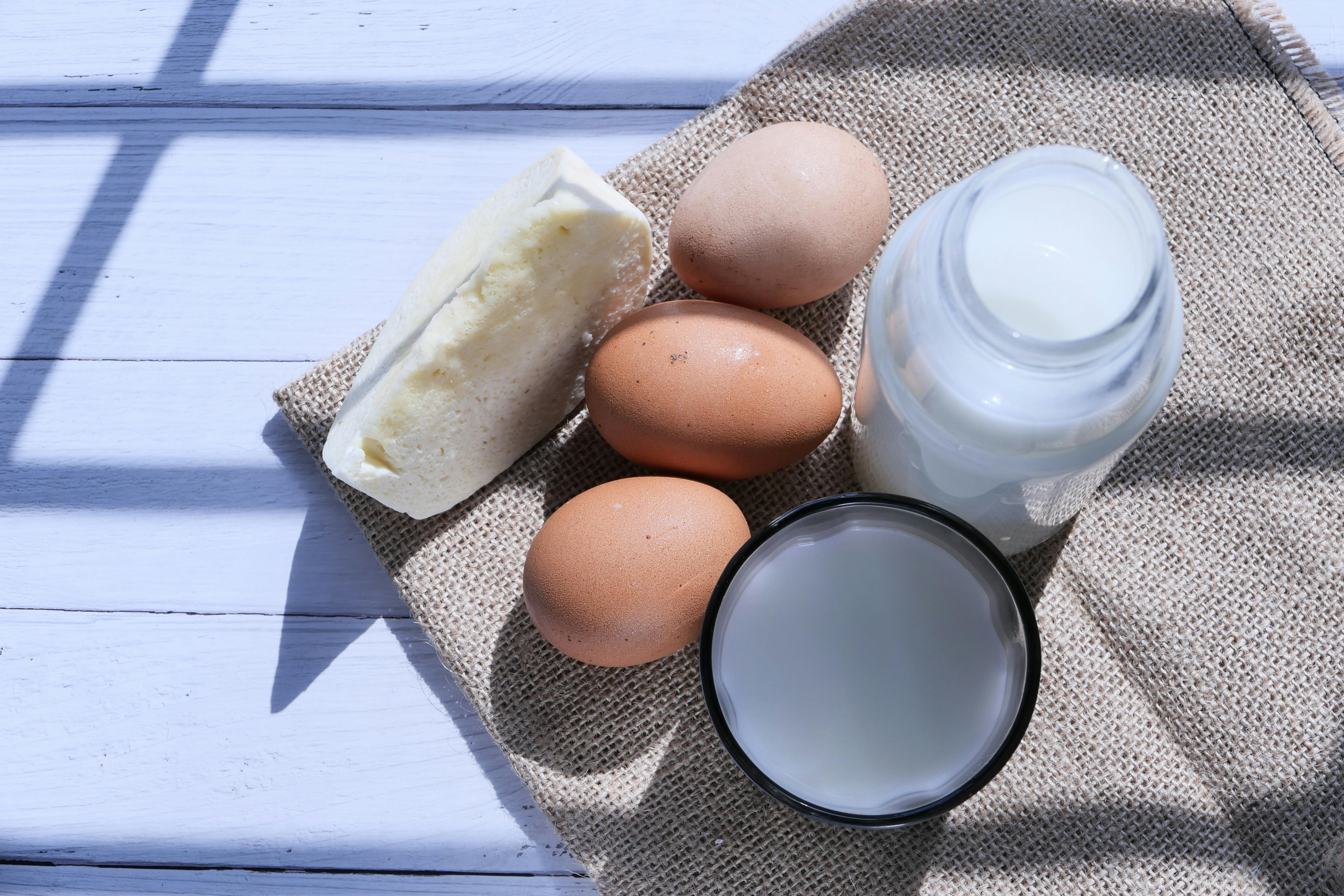 Overhead view of eggs, milk, and cheese on burlap, highlighting rustic food essentials.