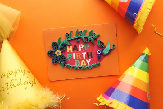 Colorful quilling birthday card surrounded by party hats on an orange backdrop, perfect for festive celebrations.