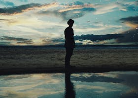A solitary figure silhouetted against a twilight beach scene with stars and reflection.