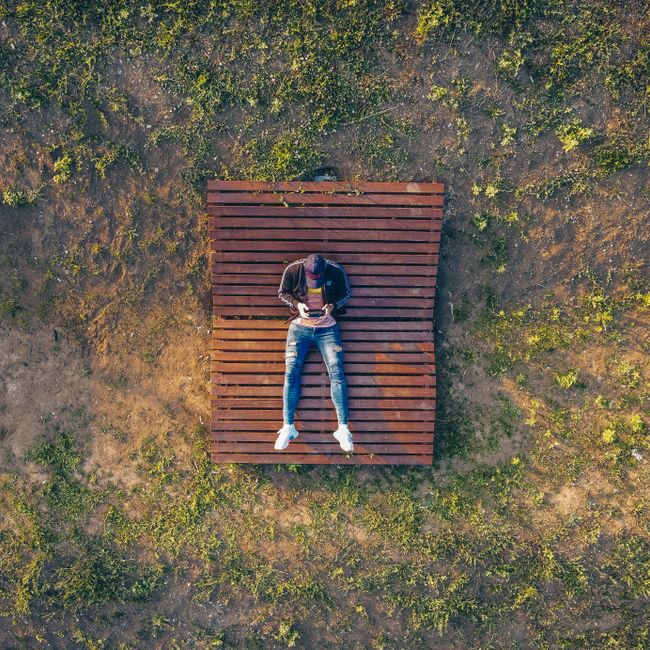 Top view aerial shot of a man relaxing on a wooden bench surrounded by grass outdoors.