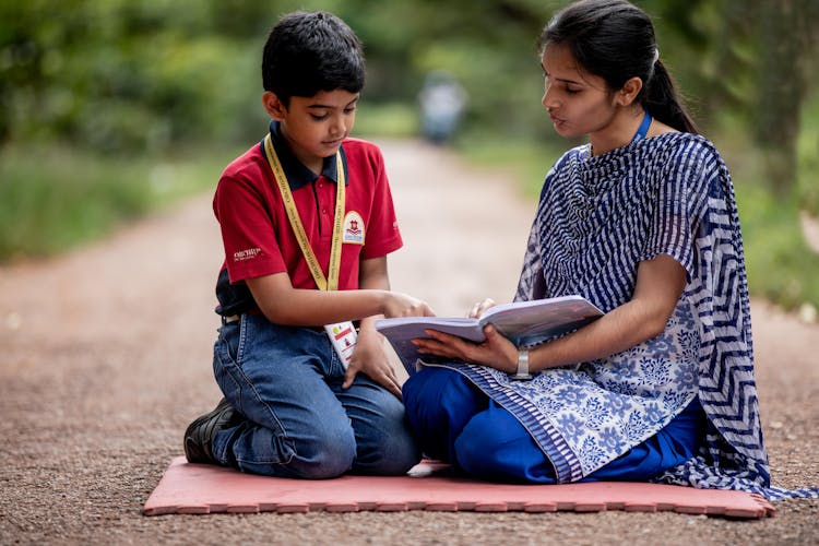 A Woman Teaching A Young Boy While Sitting On The Street