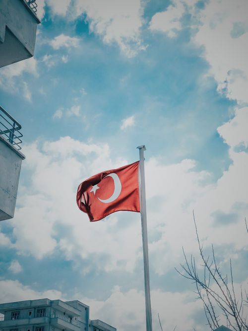Free Turkish flag waving on a flagpole with a blue sky and city buildings in the background. Stock Photo