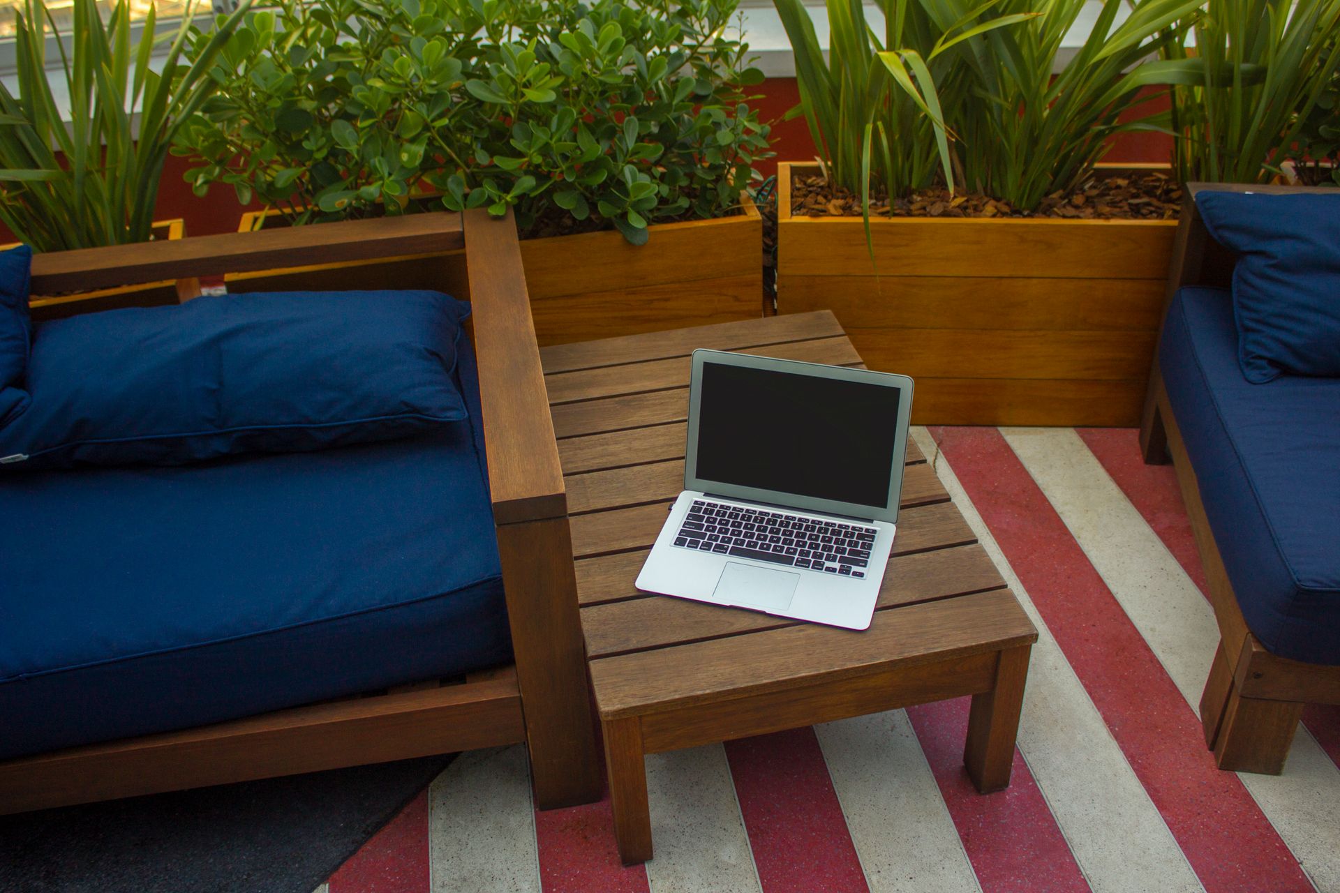 An outdoor workspace featuring a laptop on a wooden table with planters and seating area.