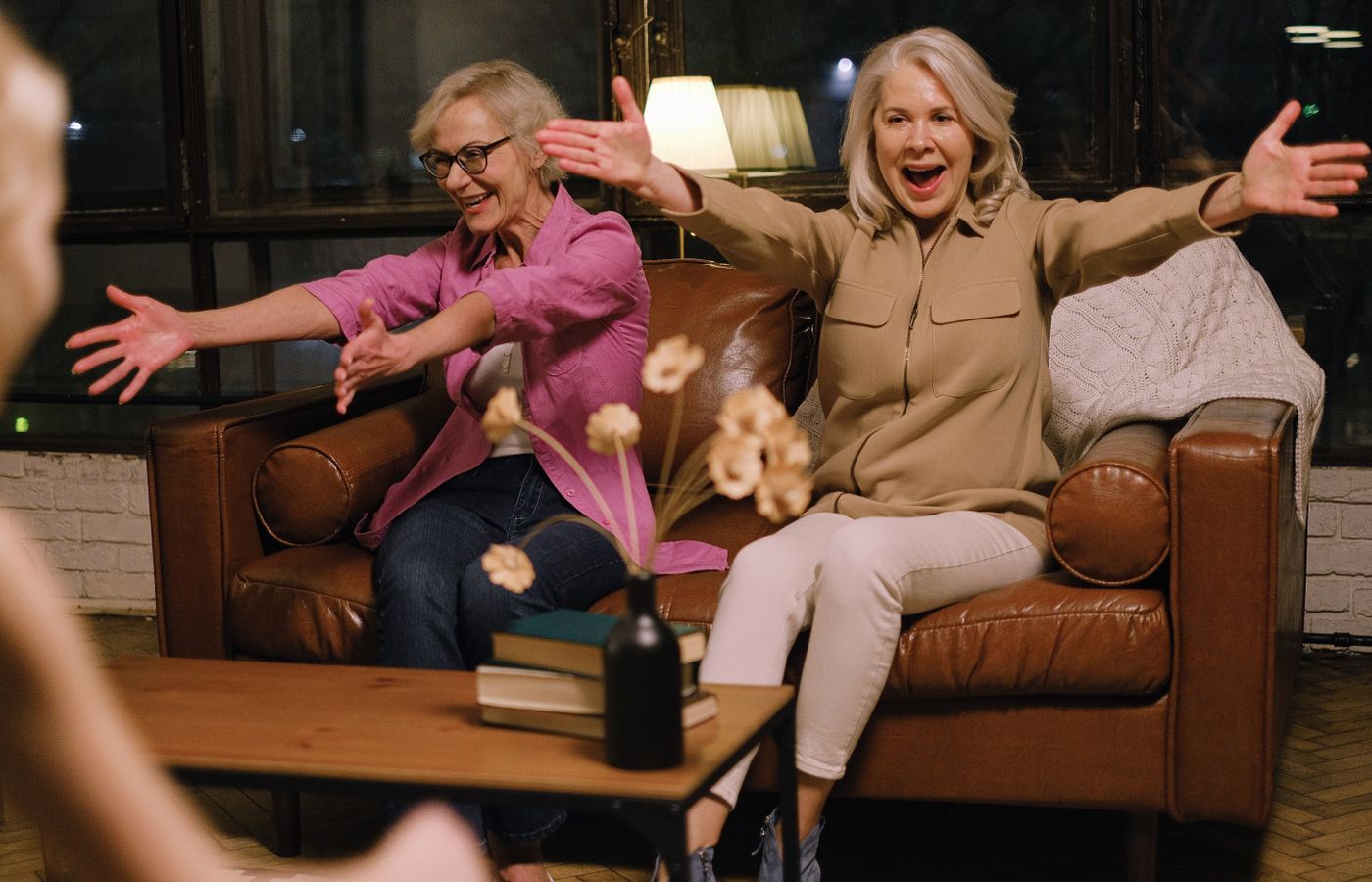 Two happy senior women on a sofa reaching out for a hug, creating a warm and joyful atmosphere indoors.