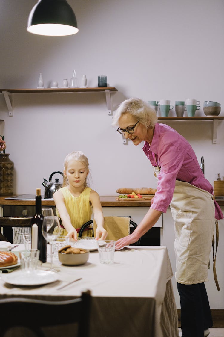 A Young Girl Helping Her Grandmother In Preparing Plates On The Table