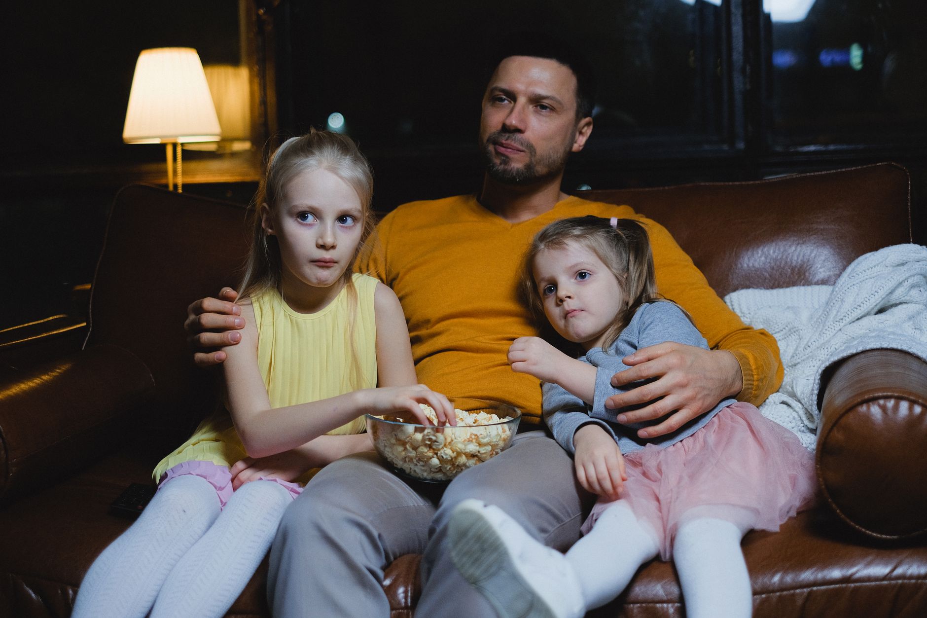 A father and his two daughters sitting on a sofa enjoying popcorn during a cozy movie night.