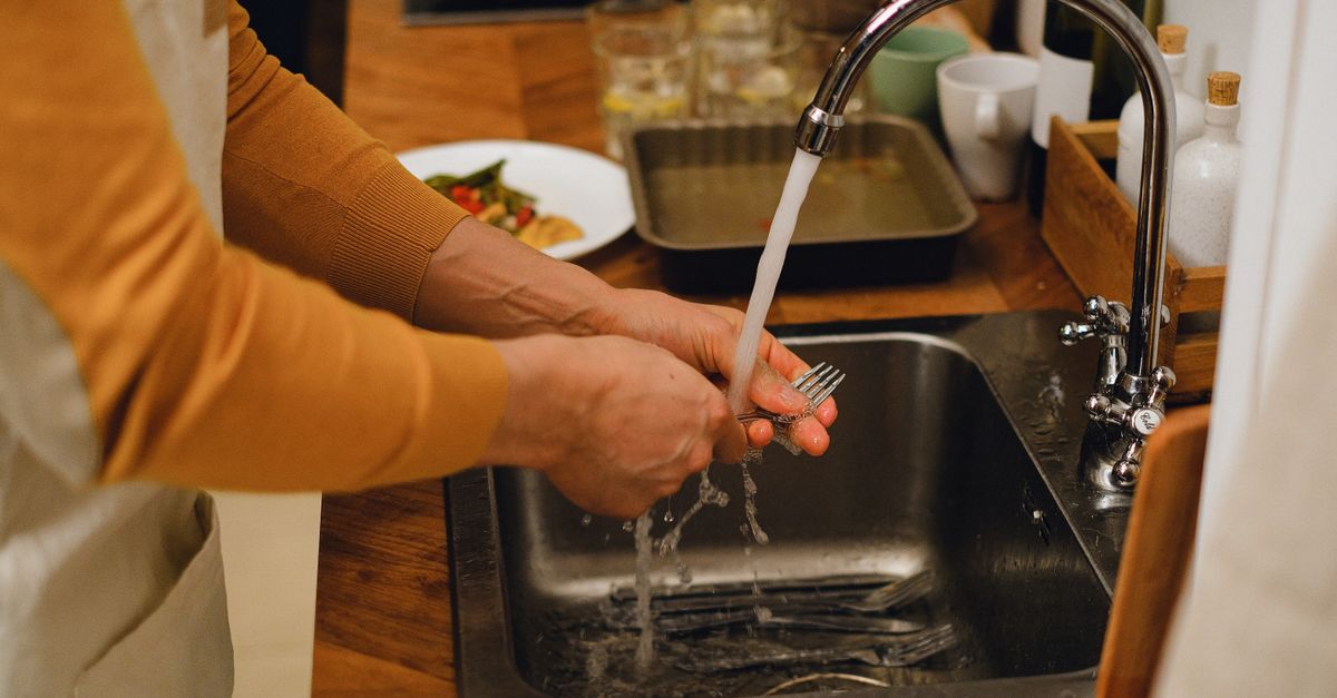 Close-up de mãos lavando um garfo sob água corrente em uma pia de cozinha com uma bancada aconchegante.