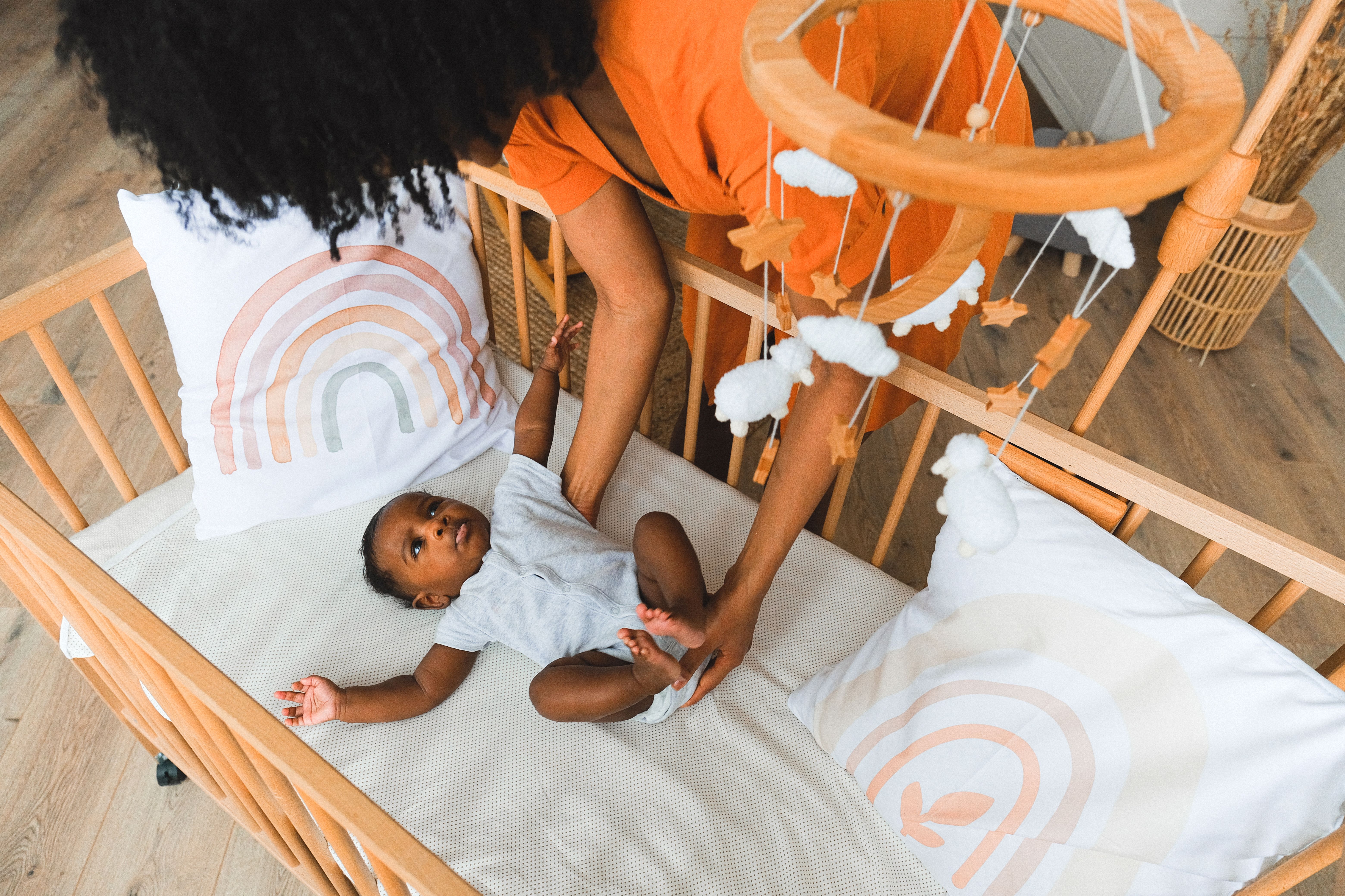 A caring mother tends to her baby in a cozy wooden crib adorned with rainbows and a whimsical mobile.