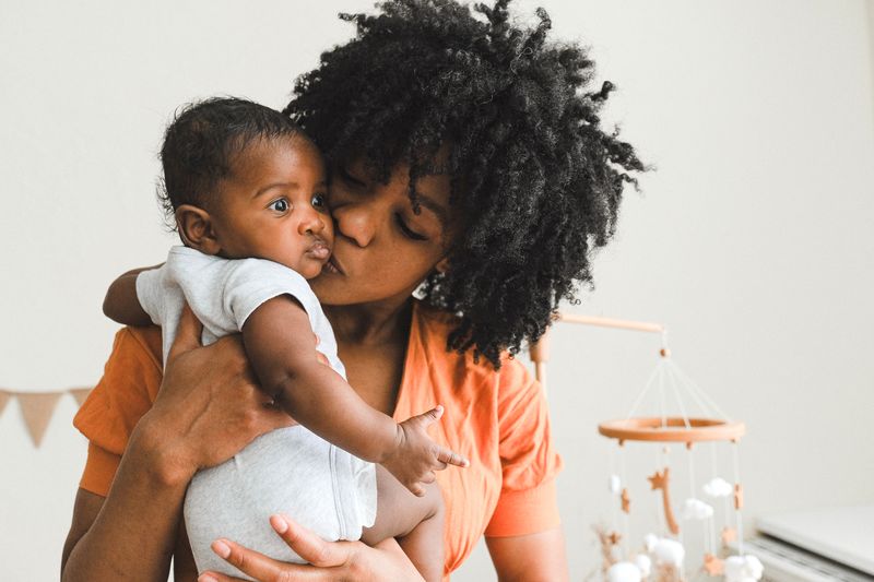 A Black mother with natural hair holds and kisses her newborn in a softly lit room, an intimate postpartum moment.
