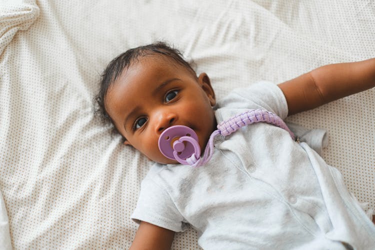 A Baby In White Onesie Lying On The Bed 