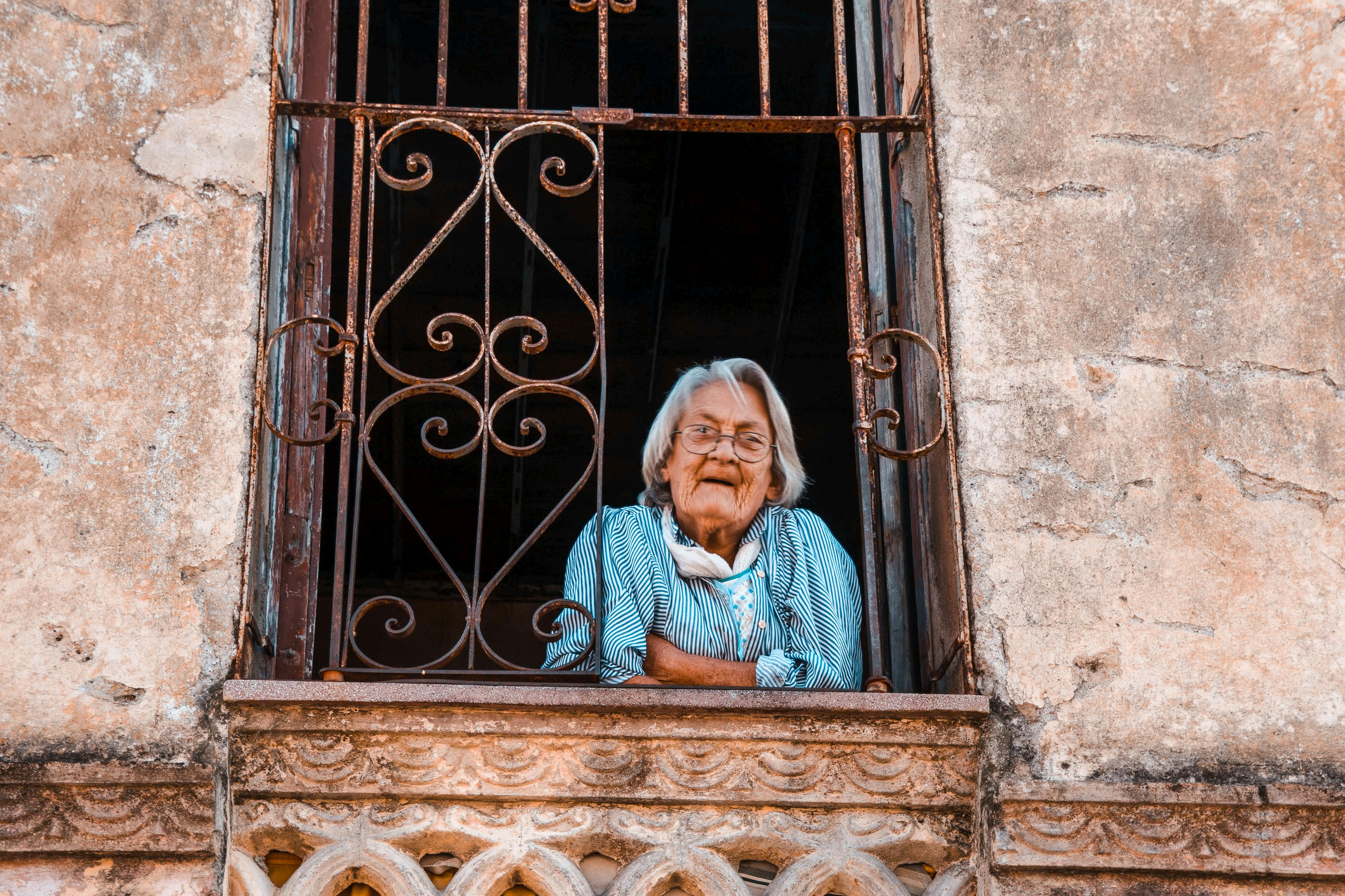 An elderly woman peers out from a rusty, ornate window of a weathered building.