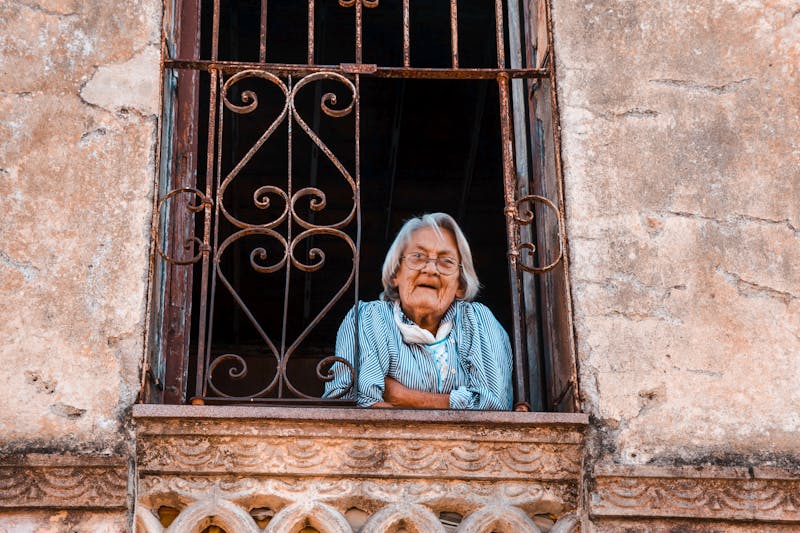 An elderly woman peers out from a rusty, ornate window of a weathered building.