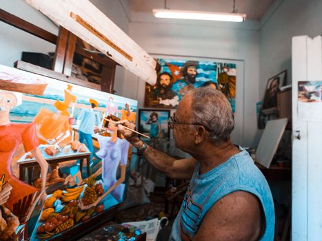 An elderly artist skillfully paints a colorful canvas in his studio, surrounded by vibrant artworks.