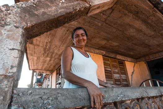 Smiling woman in white dress poses on balcony of rustic, weathered building.