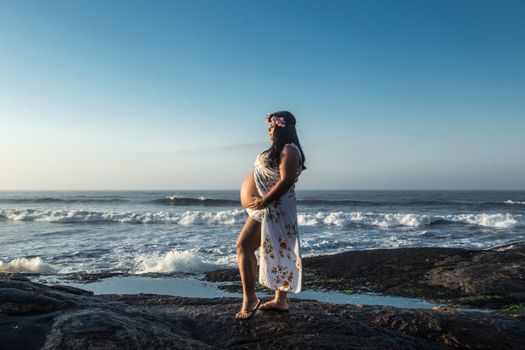 A pregnant woman stands on a rocky shore in Itanhaém, Brazil, gazing at the ocean waves.