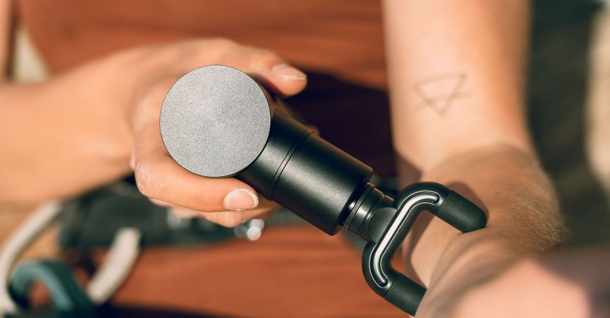 A close-up shot of a hand using a massage gun on an arm with a triangle tattoo visible.