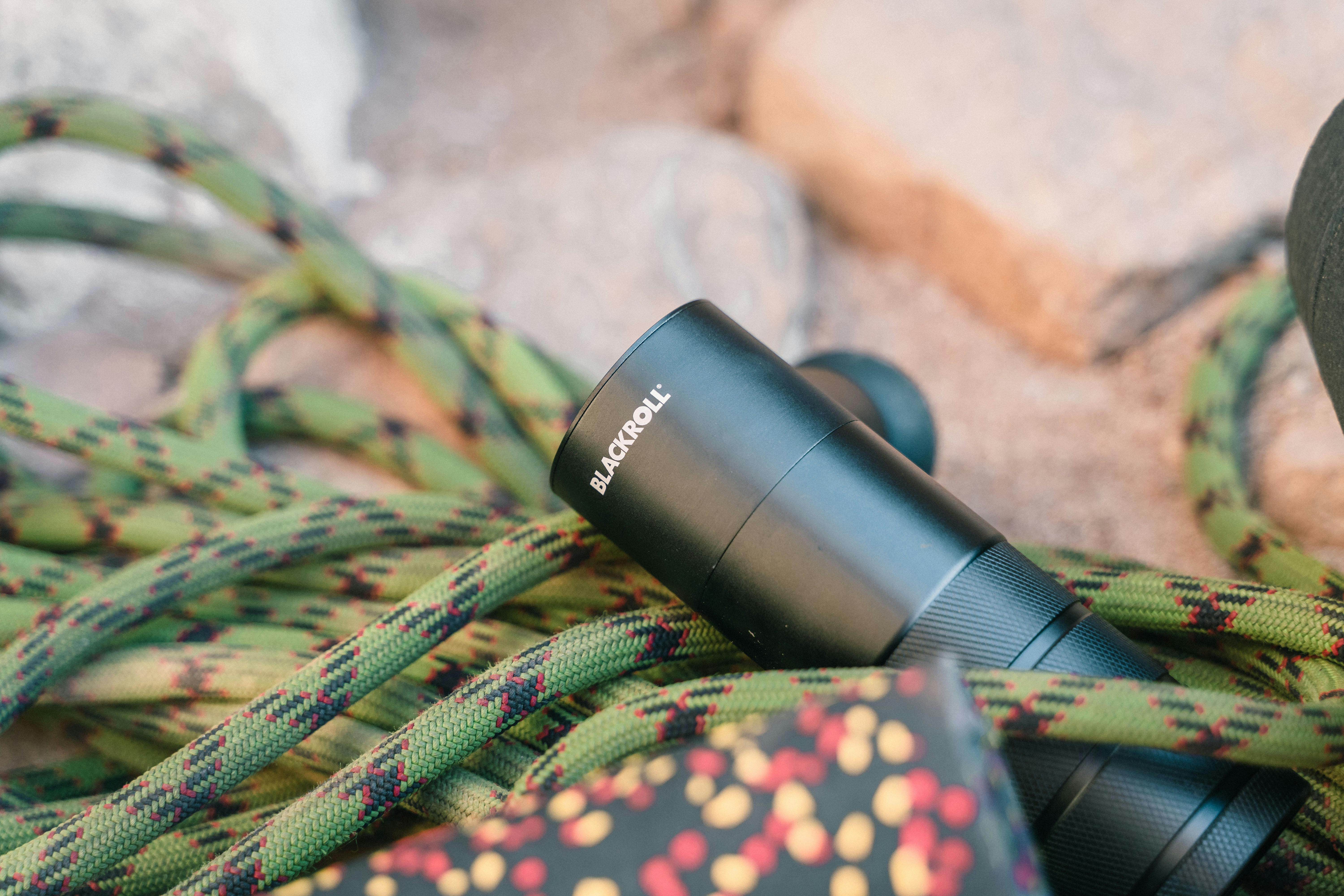 Close-up of a Blackroll massage gun and climbing rope outdoors on a sandy surface.
