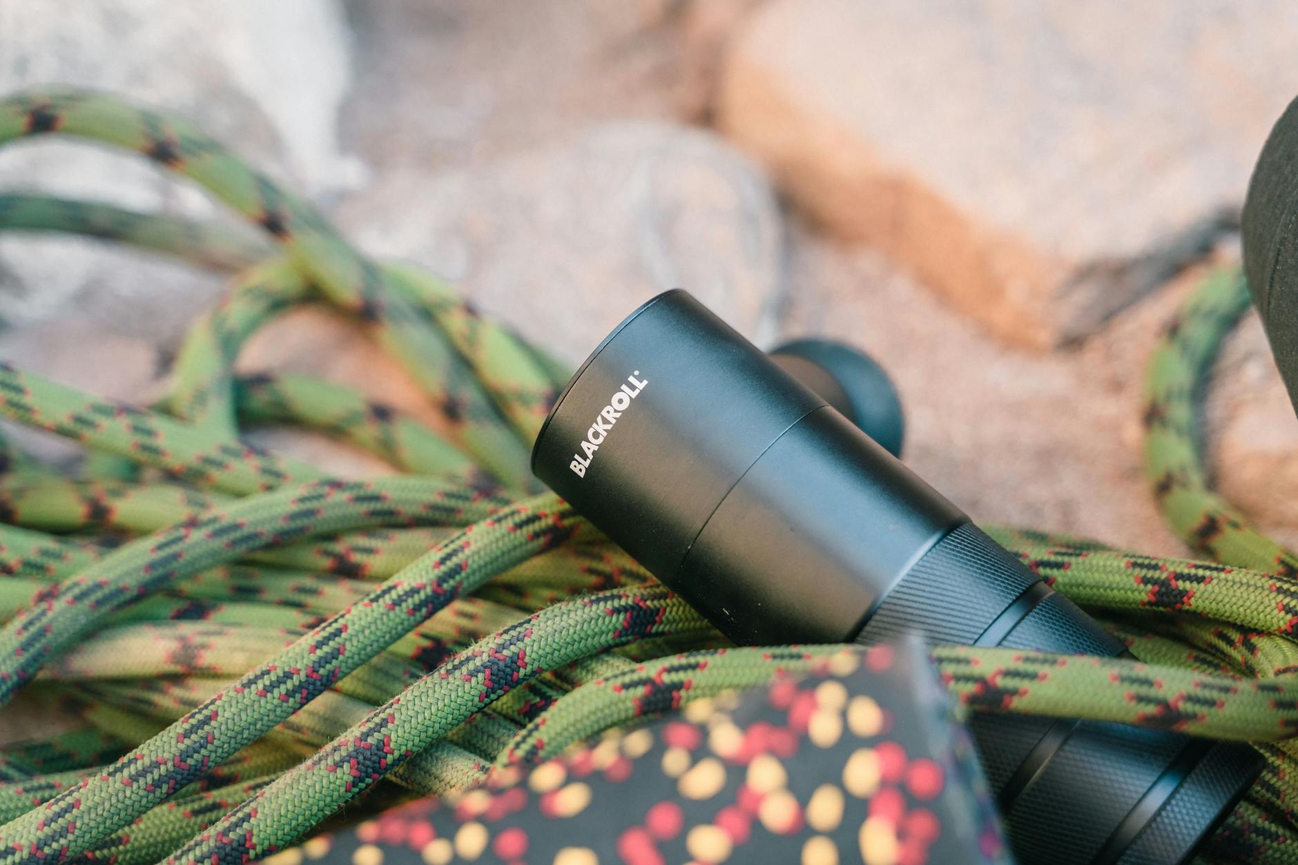 Close-up of a Blackroll massage gun and climbing rope outdoors on a sandy surface.
