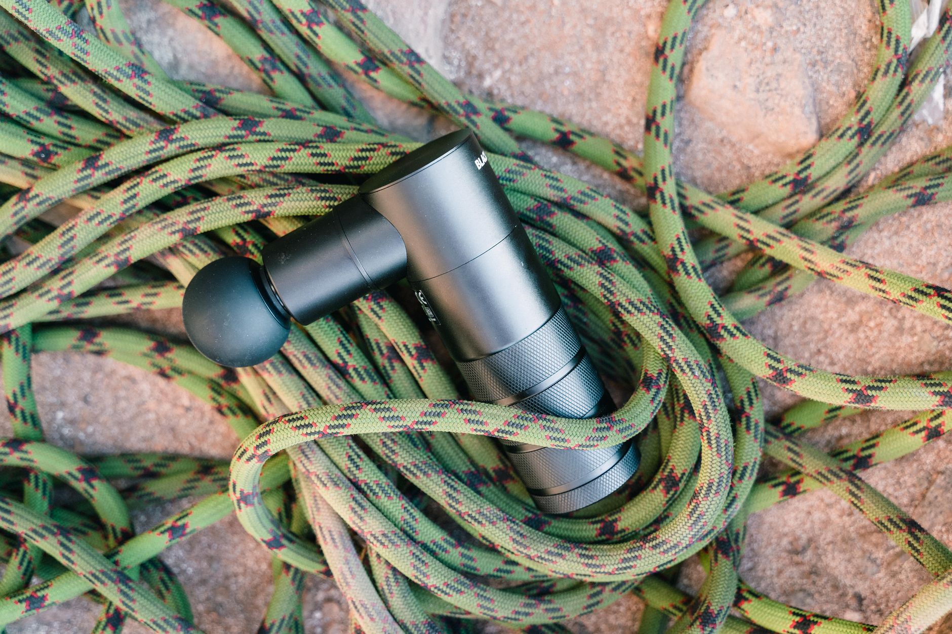 A massage gun laying on a coiled climbing rope on a rocky surface, showcasing fitness and adventure tools.