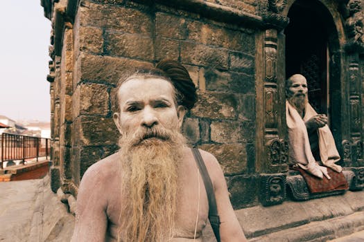 Two traditional South Asian holy men with beards sitting outside a historic stone building.