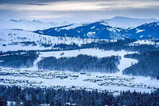 Scenic winter view of a snowy village nestled in a mountainous landscape with forested hills.