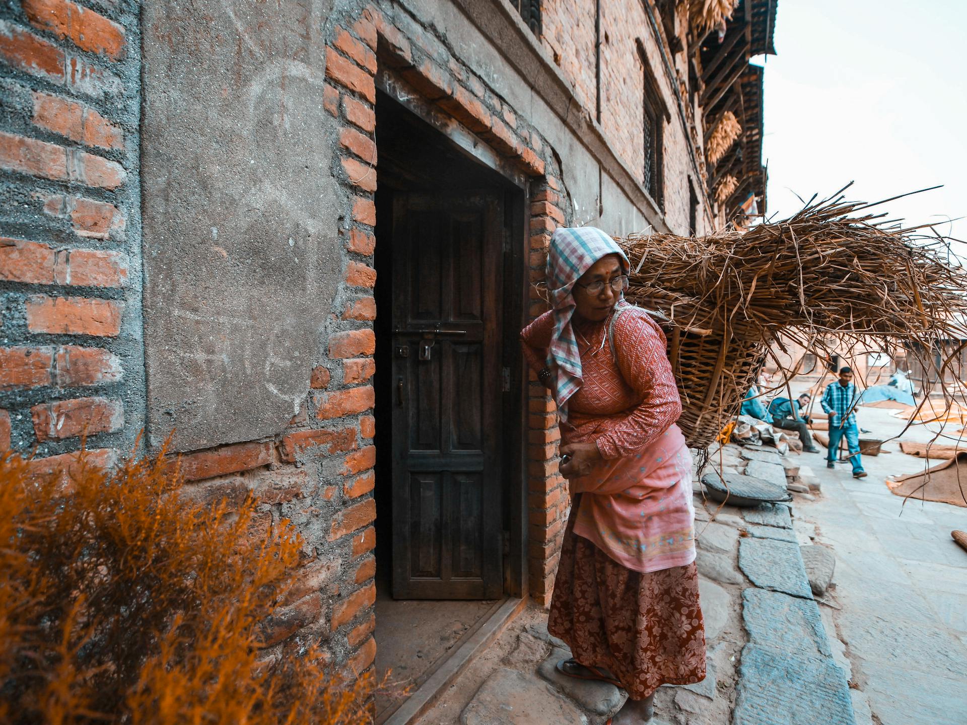 A village woman carrying a brushwood basket enters a rustic building.