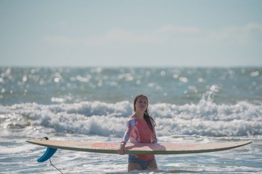 Caucasian woman in a rash guard stands with a surfboard in ocean waves on a sunny day.