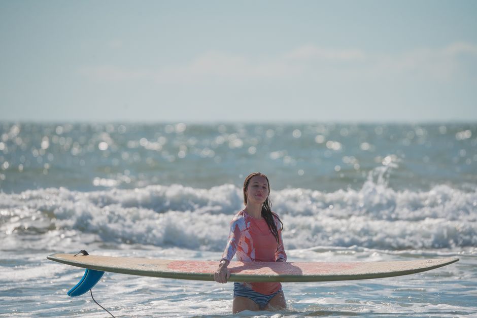 Caucasian woman in a rash guard stands with a surfboard in ocean waves on a sunny day.