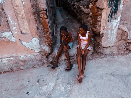 Free Two girls sitting on a rustic alley floor, surrounded by aged stone walls. Stock Photo