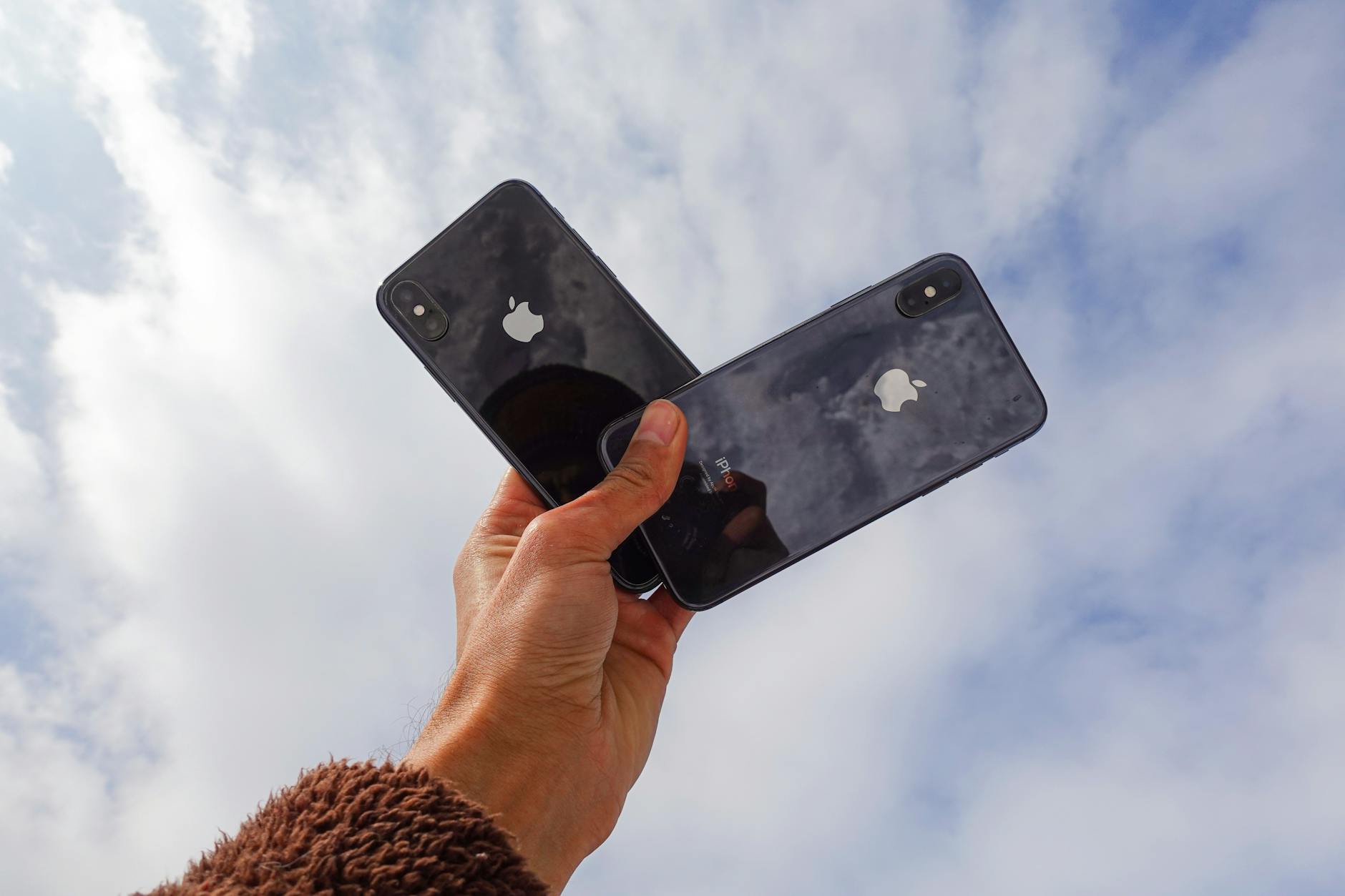 Close-up of a hand holding two smartphones against a cloudy sky, showcasing modern technology. Apple Refurbished vs. Used iPhones: Which Gives You More Value