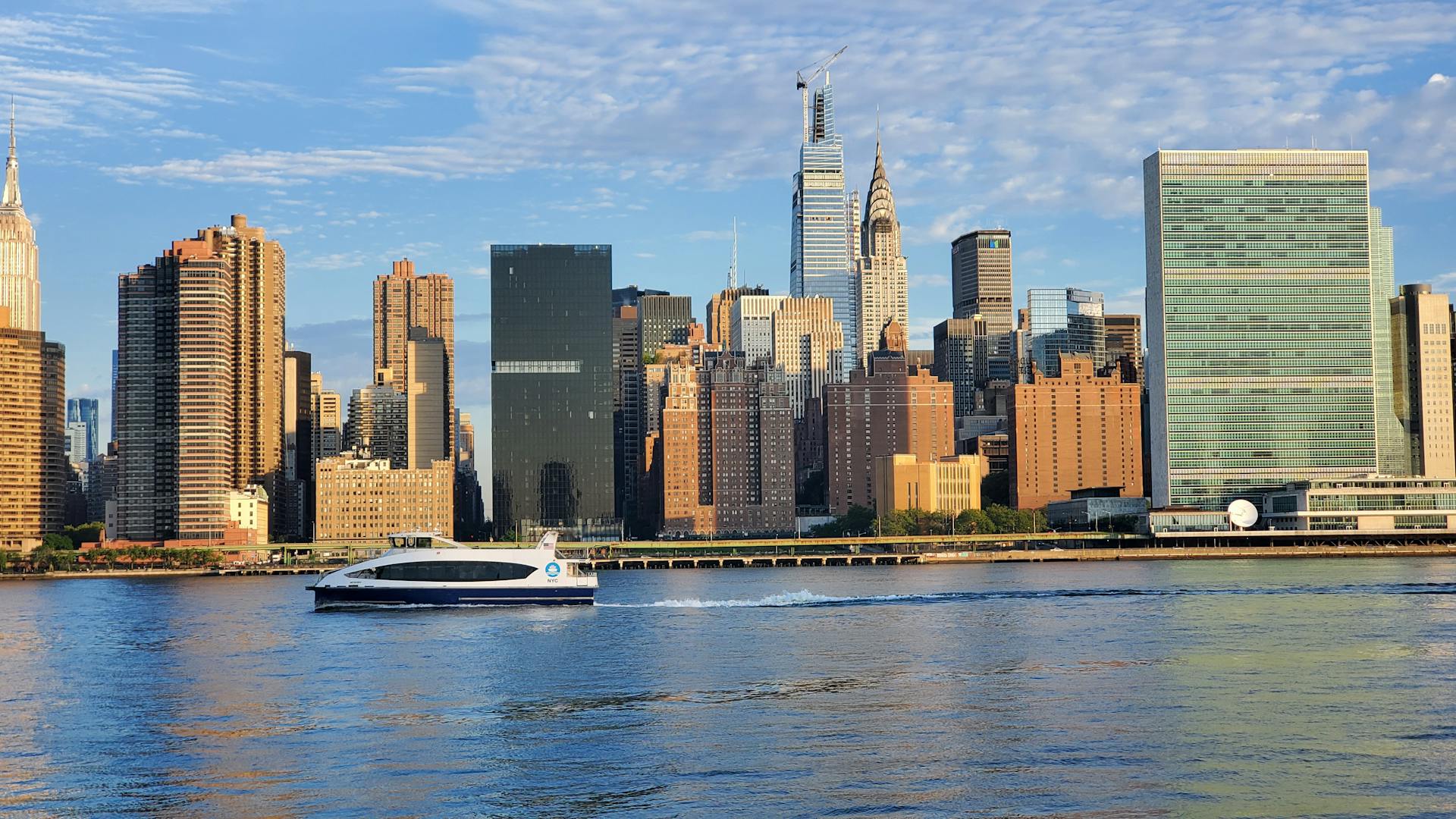 A ferry boat cruises along the waterfront with the iconic New York City skyline in the background.