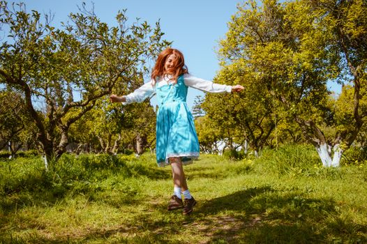 A cheerful young girl jumps with joy in a lush, sunlit orchard with green trees and grass.