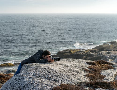 Man in Black Jacket Leaning on Rock Near Sea Taking a Photo