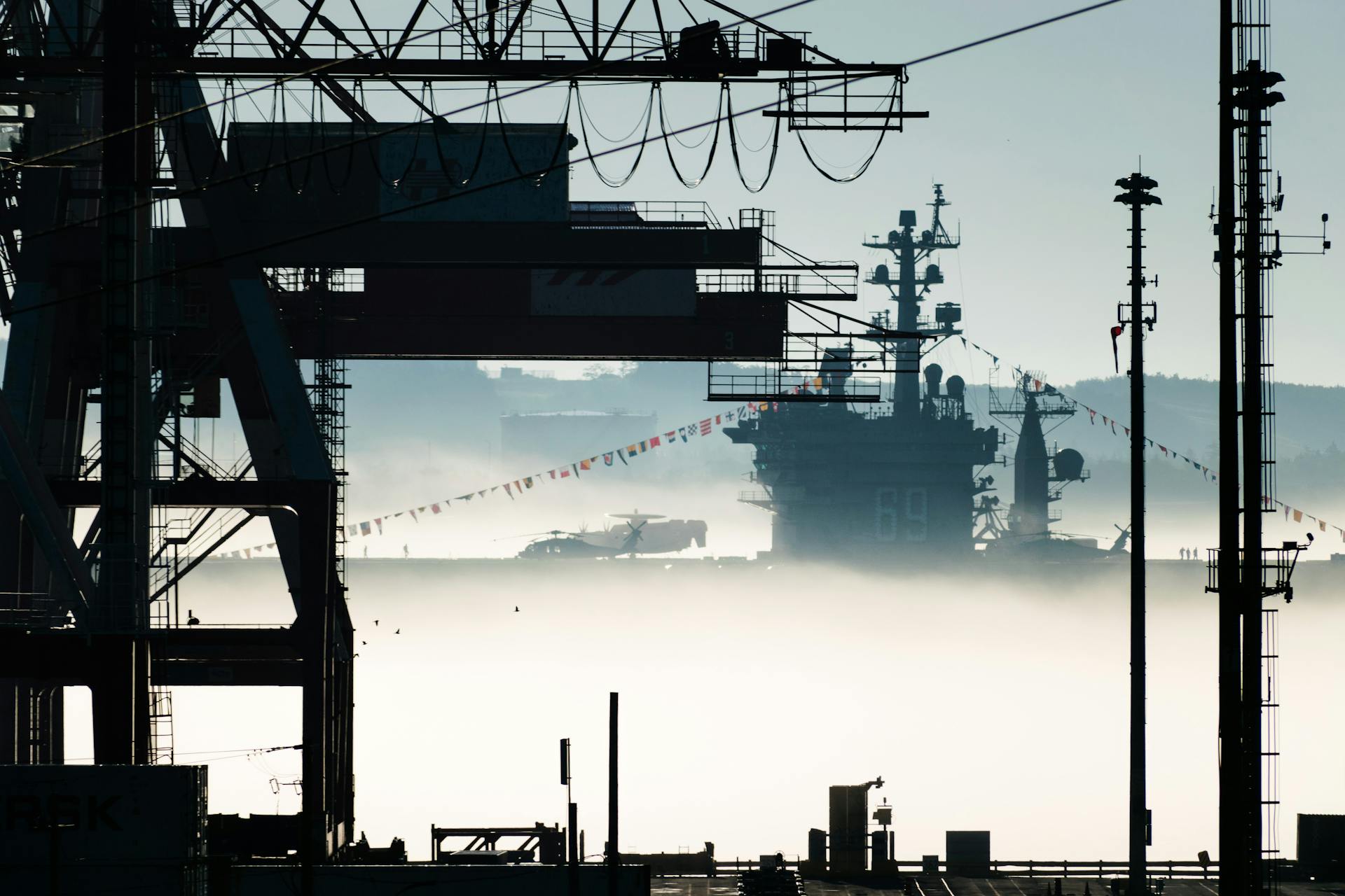 Military ship in a misty harbor with cranes and fog, showcasing maritime infrastructure.