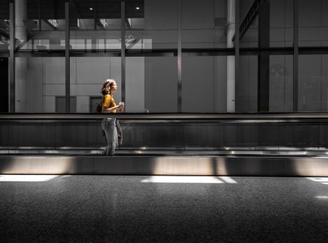 Side view of a woman walking through an airy Toronto airport terminal with a plastic cup in hand.