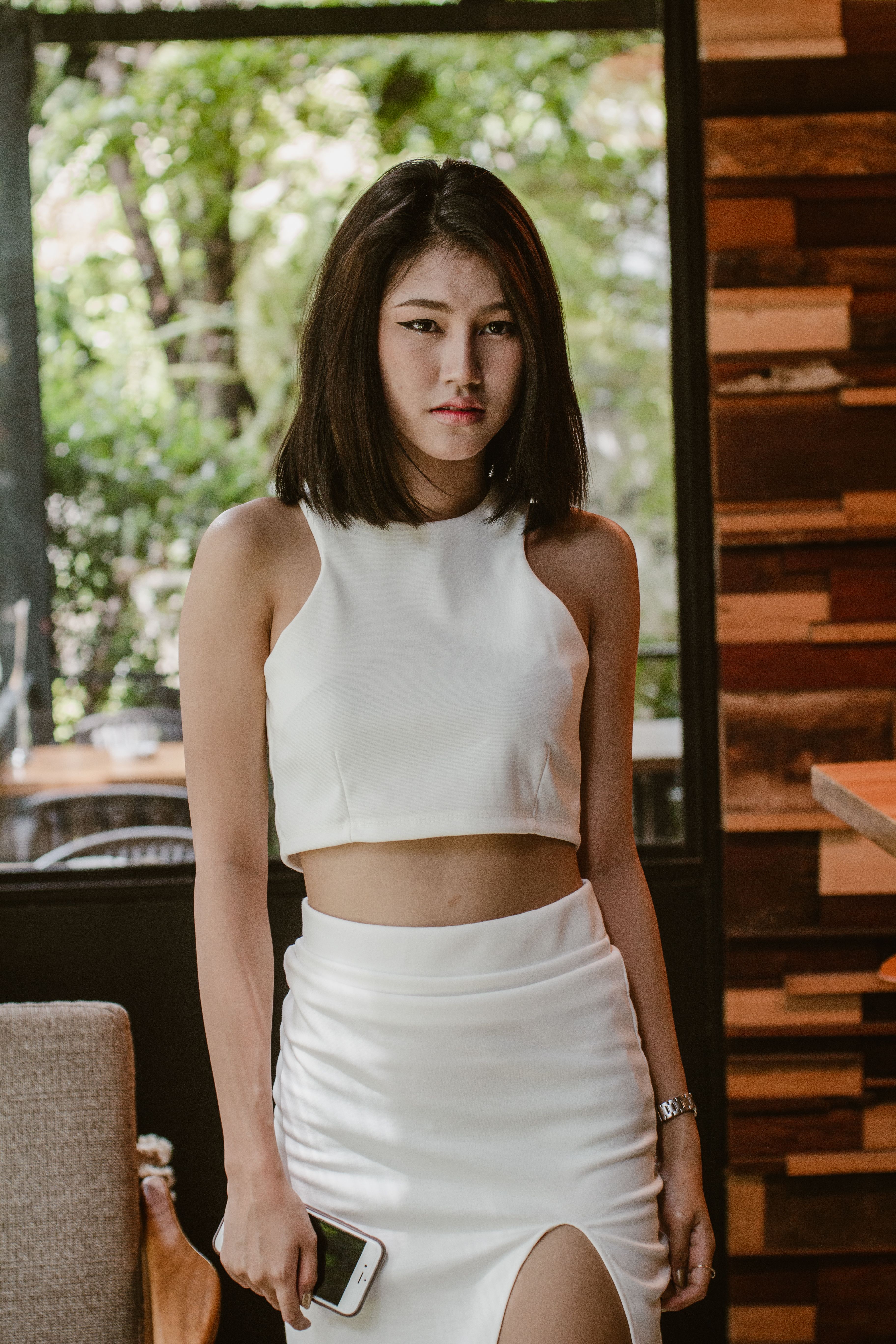 Stylish young woman wearing a white crop top and skirt, holding a smartphone indoors.