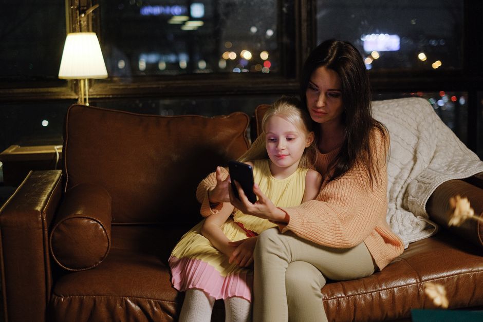 Photo by Anna Shvets on Pexels A mother and daughter share quality time on a leather couch, illuminated by a cozy lamp, with a city nightscape in the background. - Photo by Anna Shvets on Pexels